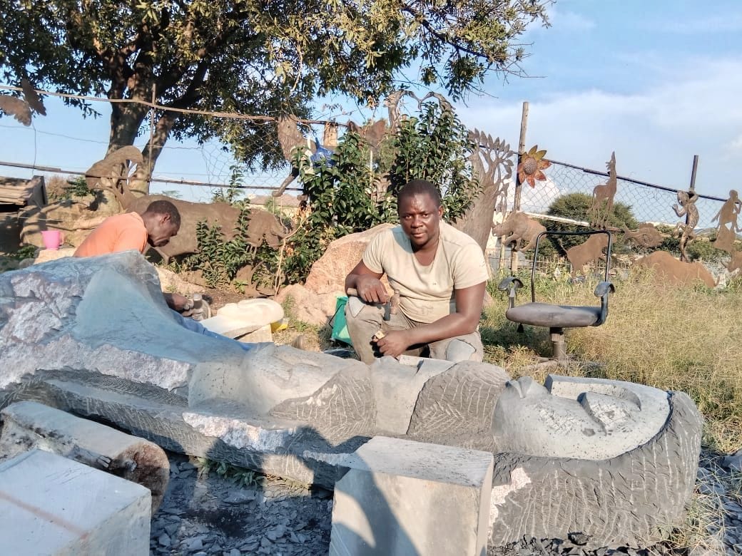 A carver in a beige T-shirt seated beside the stone in a workshop yard