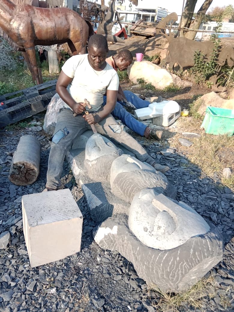 A carver shaping the defined faces of the work with a block of stone beside him