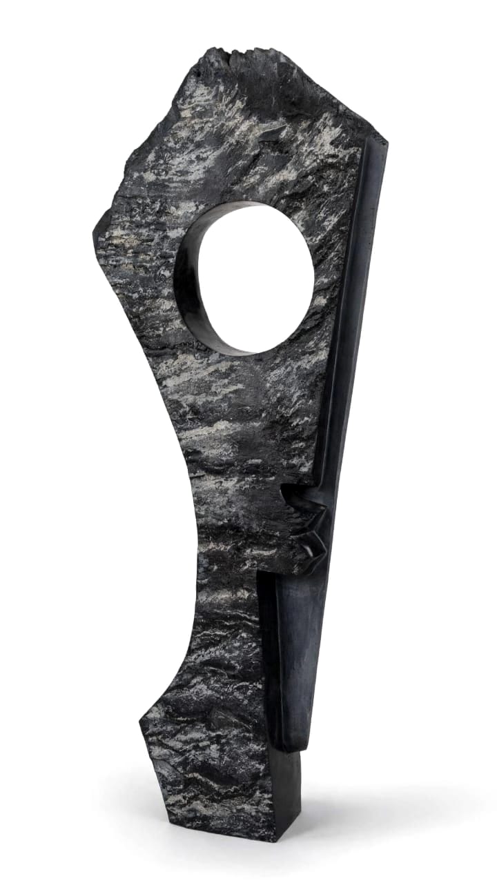 Tall grey-black stone sculpture with a round opening and a dark carved base, on white