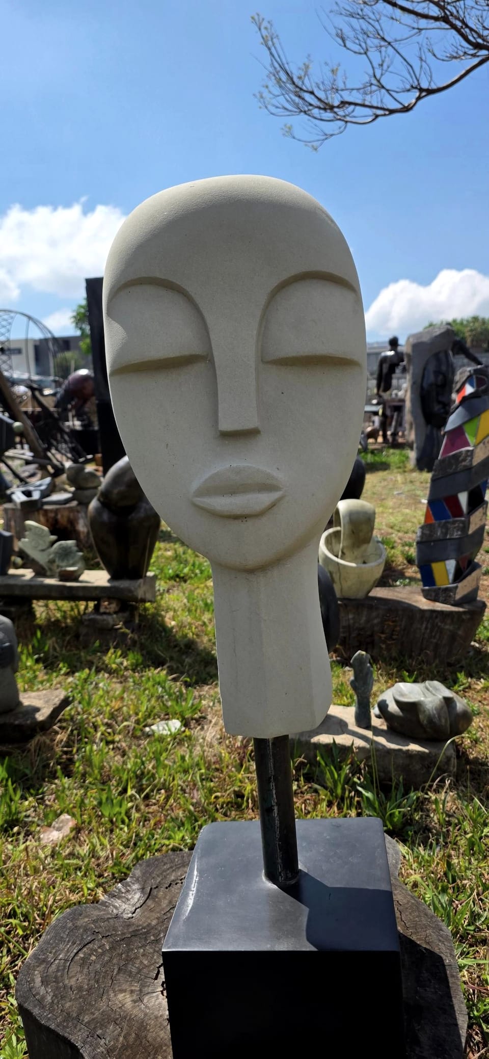 Pale stone head with closed eyes and full lips, raised on a dark metal stem and square black base, in the workshop yard