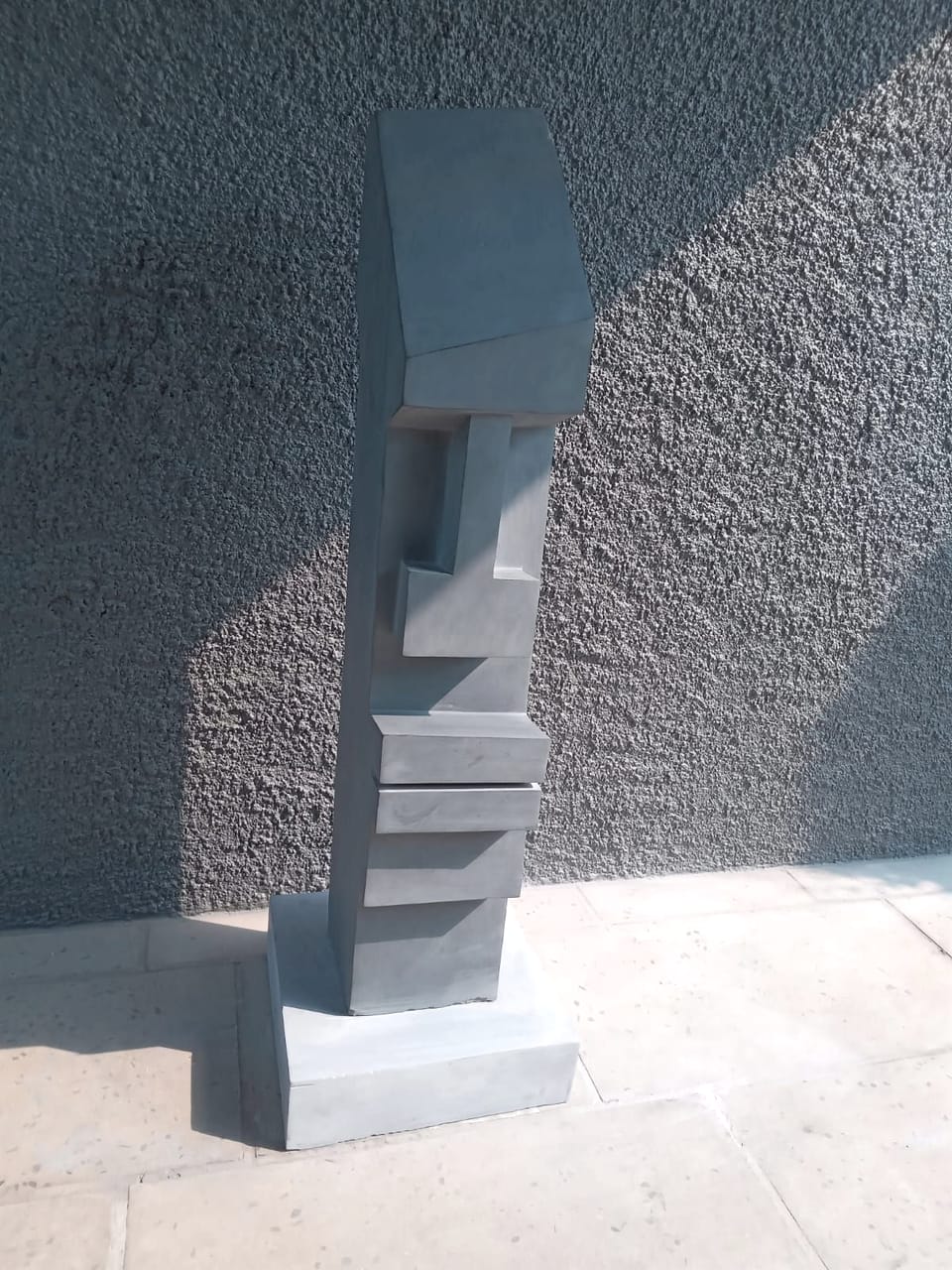 A grey stone column with an angled crown, a cut-in eye and stacked blocks for the mouth, standing on a square base against a textured wall