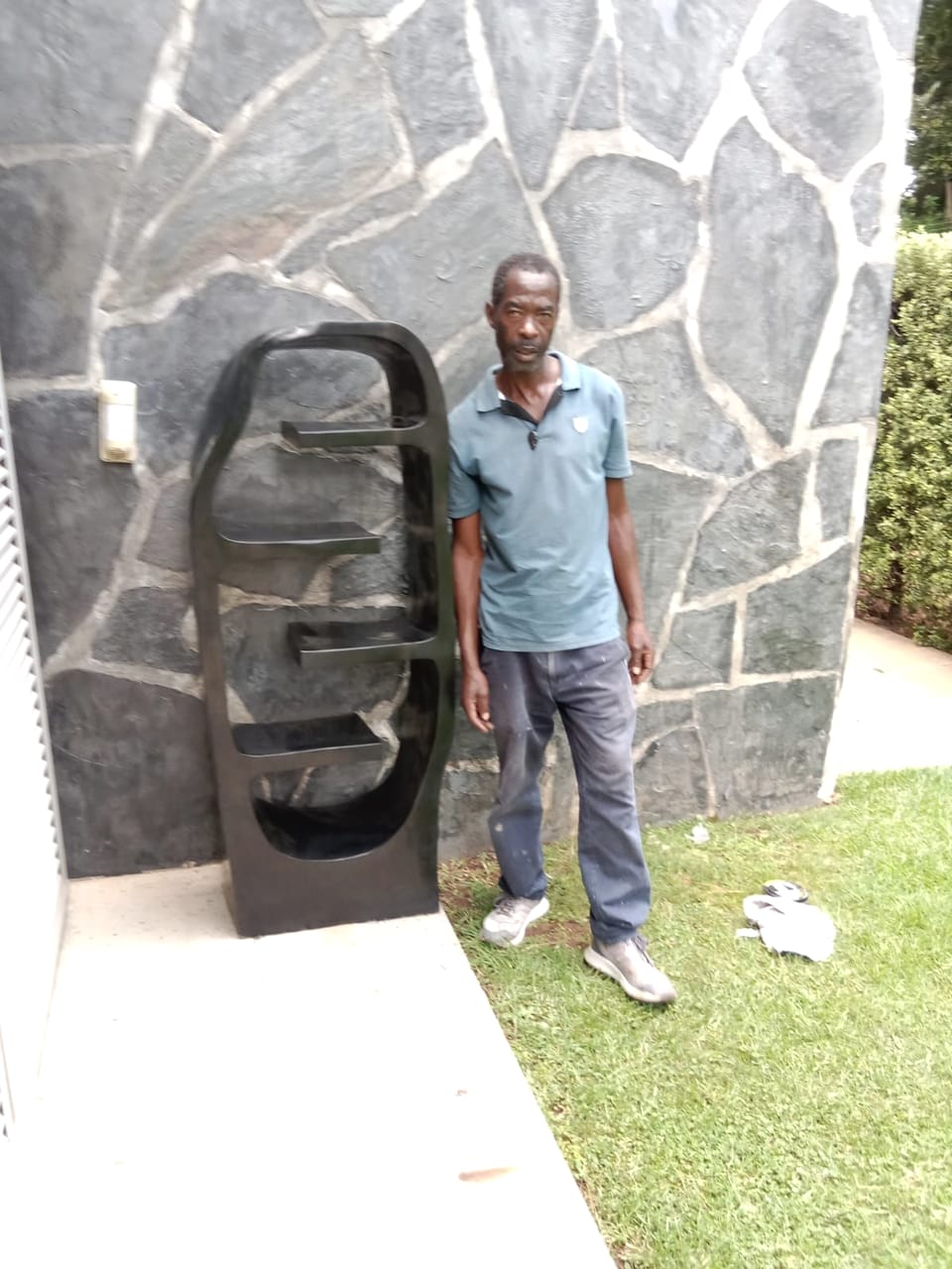 Tall dark stone shelf with four ledges set inside an open, rounded frame, Nomore standing beside it in a garden