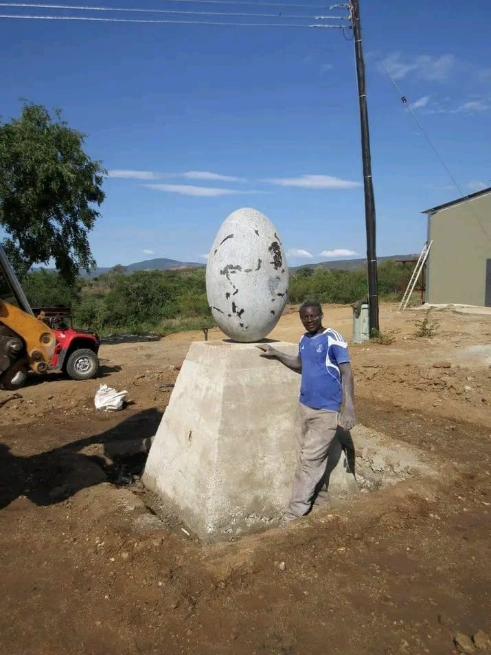 Pale egg-shaped sculpture on a stone plinth, with a man beside it in the yard