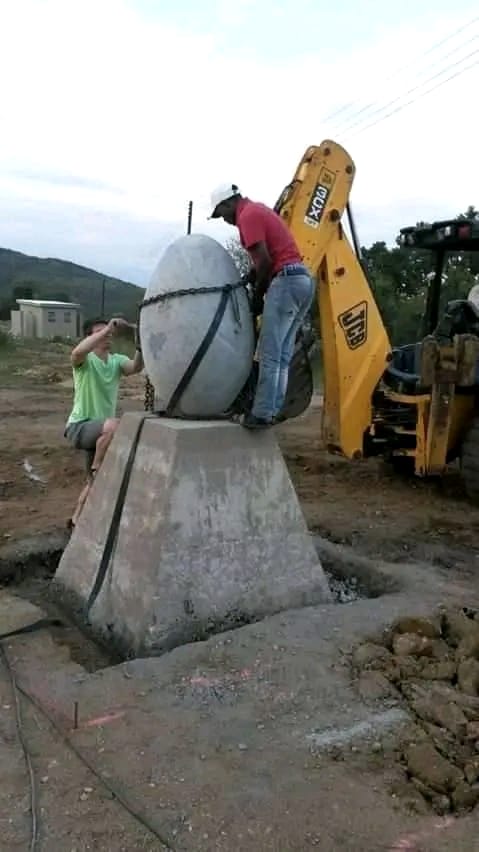 Egg-shaped sculpture being lifted onto its stone plinth by chains and a digger