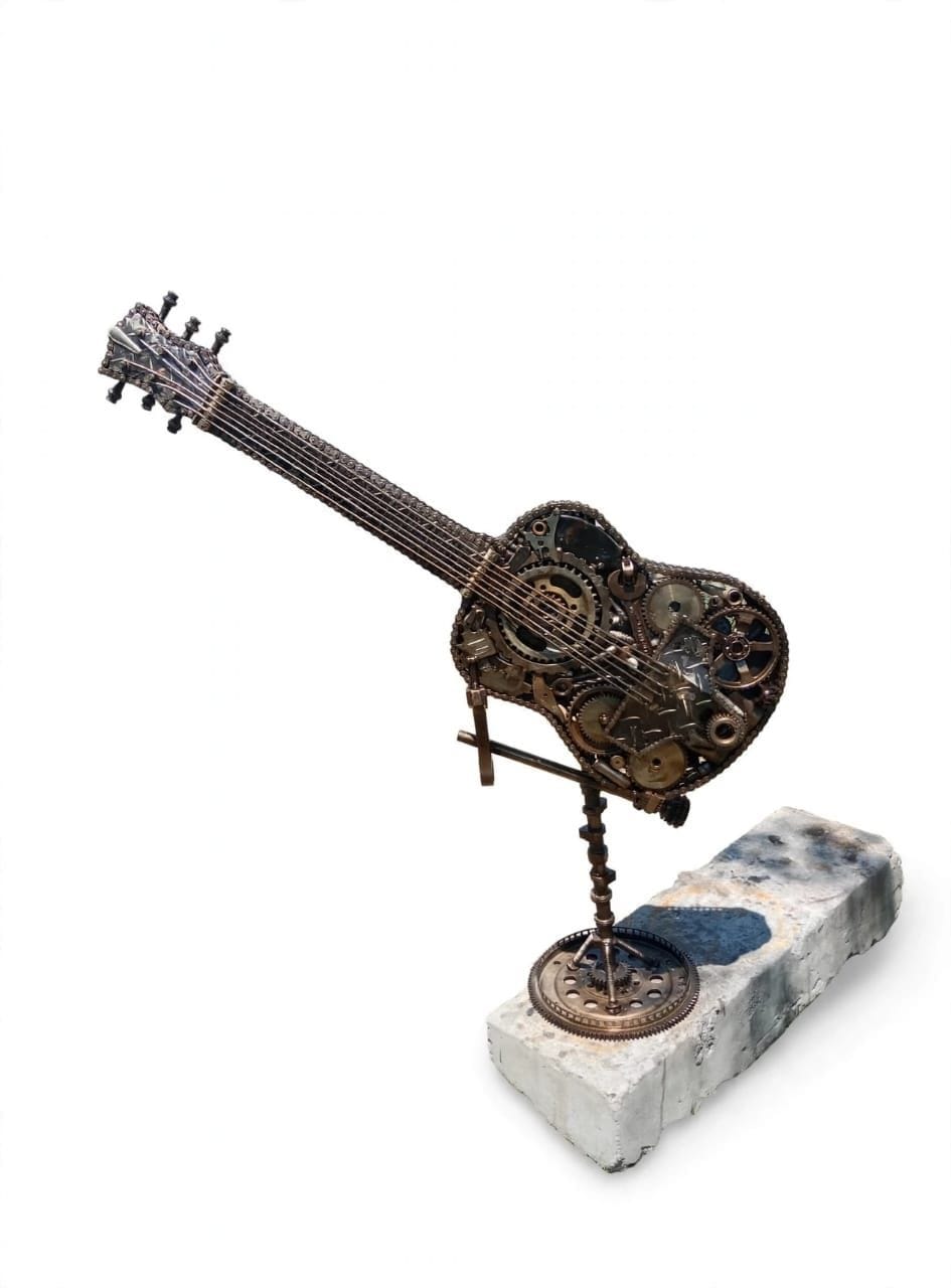 Welded metal acoustic guitar built from gears and found objects, mounted on a pale stone block, on white