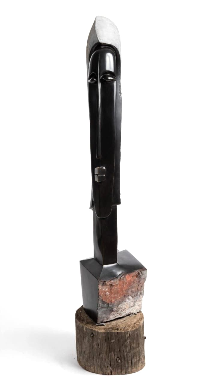 Slender polished dark stone column with an arched head, rising from a rust-coloured block on a wooden base